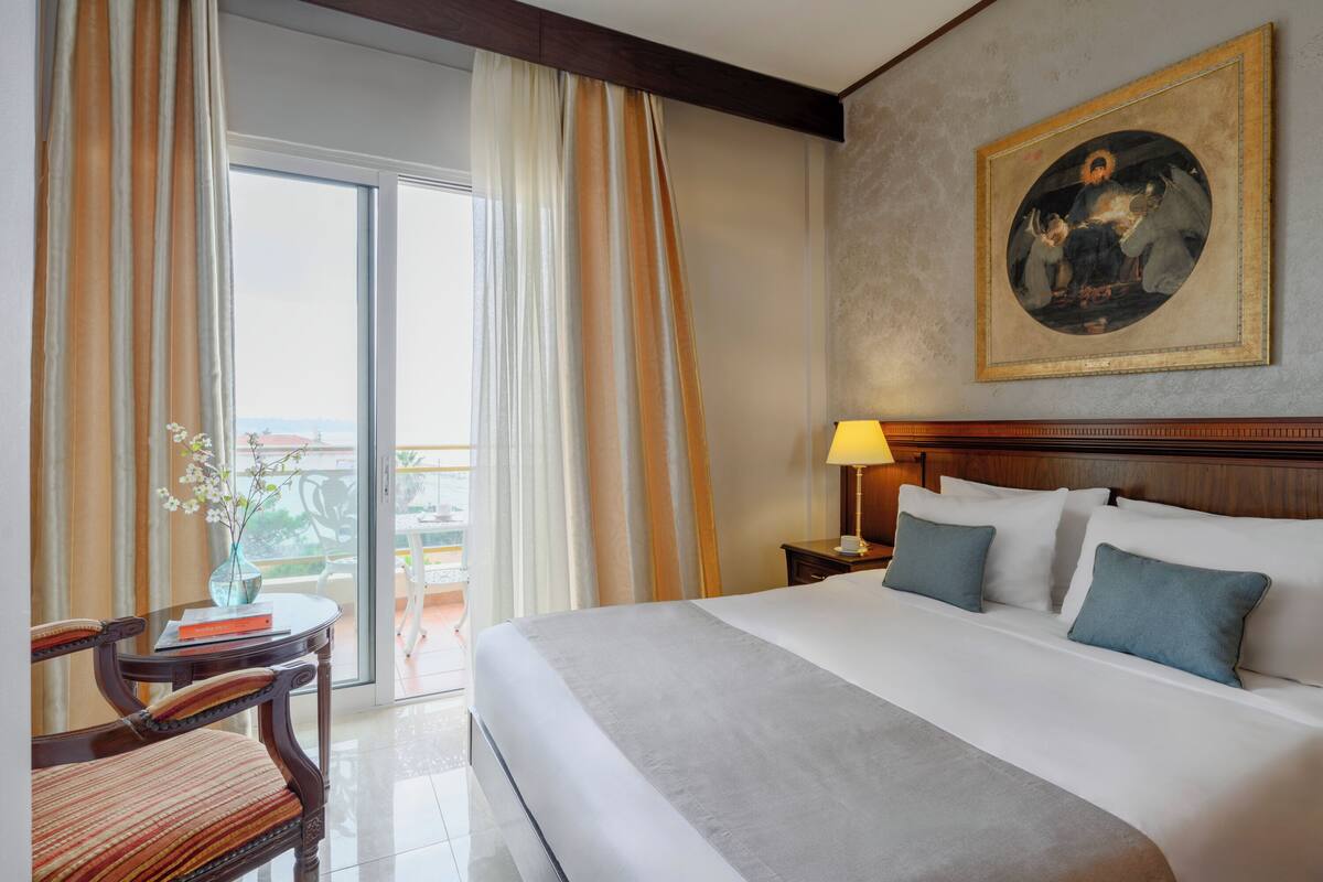 A hotel room with a large bed, a nightstand with a lamp, a chair, a table with a vase, and a painting on the wall.