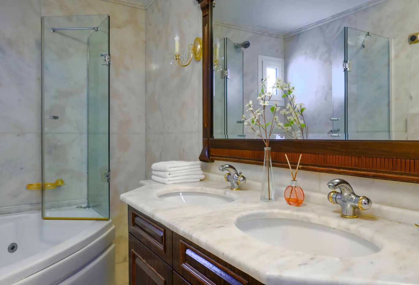 A bathroom with two sinks, a glass shower enclosure, a large mirror, and a vase with flowers.