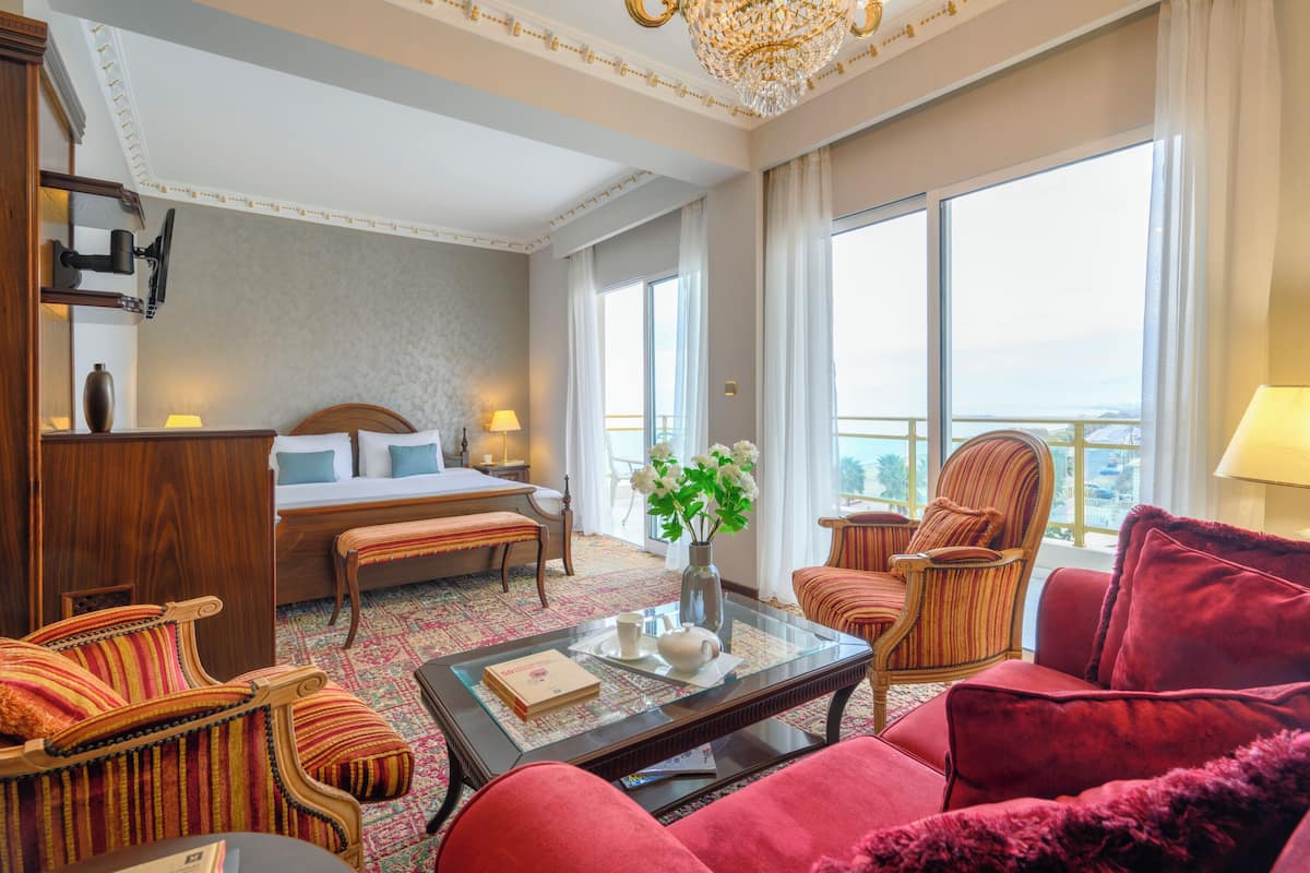 A hotel room with a bed, a sitting area with a red sofa and armchairs, a coffee table, and a view of the city through large windows.