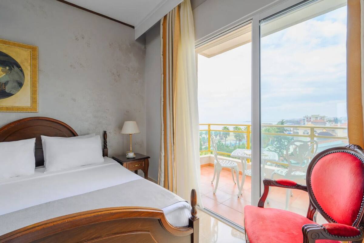 A hotel room with a bed, a nightstand, a lamp, a red chair, and a balcony with a view of the city.