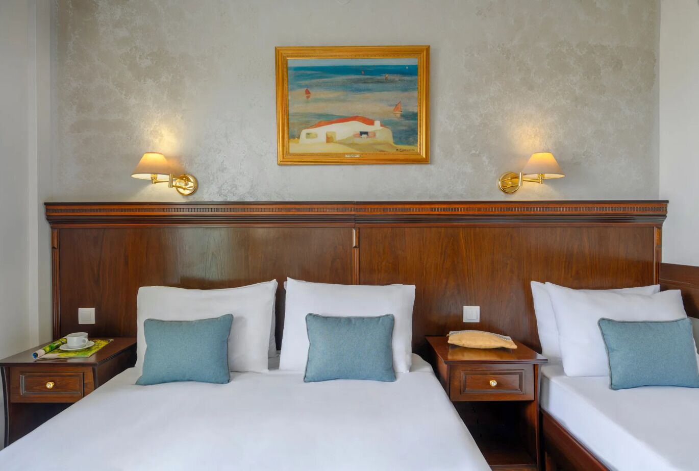 A hotel room with two beds, each with white linens and blue pillows, wooden headboards, and bedside tables with lamps. A painting of a coastal scene hangs on the wall above the beds.