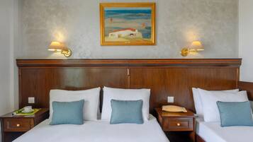 A hotel room with two beds, each with white linens and blue pillows, wooden headboards, and bedside tables with lamps. A painting of a coastal scene hangs on the wall above the beds.