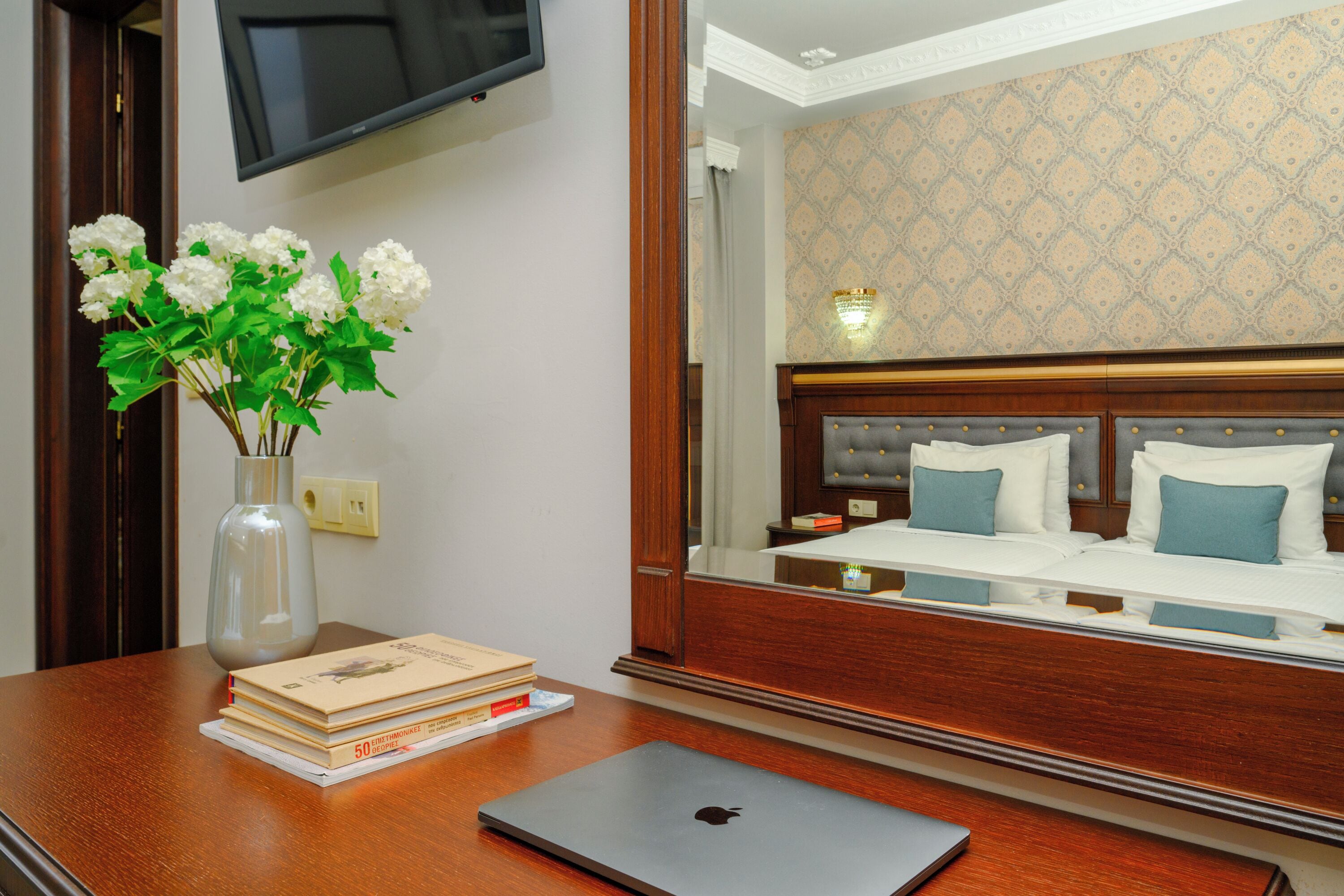 A hotel room with a wooden desk, a laptop, a vase with flowers, and a bed with blue pillows.