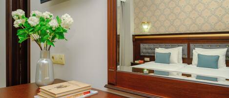 A hotel room with a wooden desk, a laptop, a vase with flowers, and a bed with blue pillows.