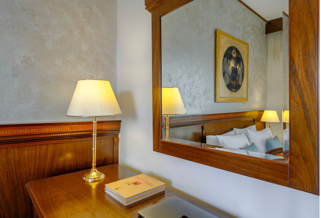 Quarto de hotel com cabeceira de madeira, mesa de cabeceira com abajur, espelho refletindo a cama e uma pintura, e uma pilha de livros.