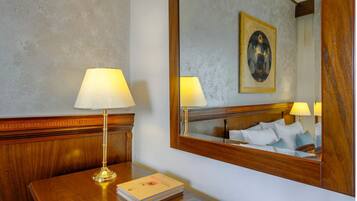 Quarto de hotel com cabeceira de madeira, mesa de cabeceira com abajur, espelho refletindo a cama e uma pintura, e uma pilha de livros.