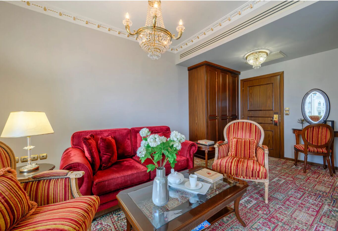Un salon avec un canapé rouge, des fauteuils à rayures, une table basse et un lustre.