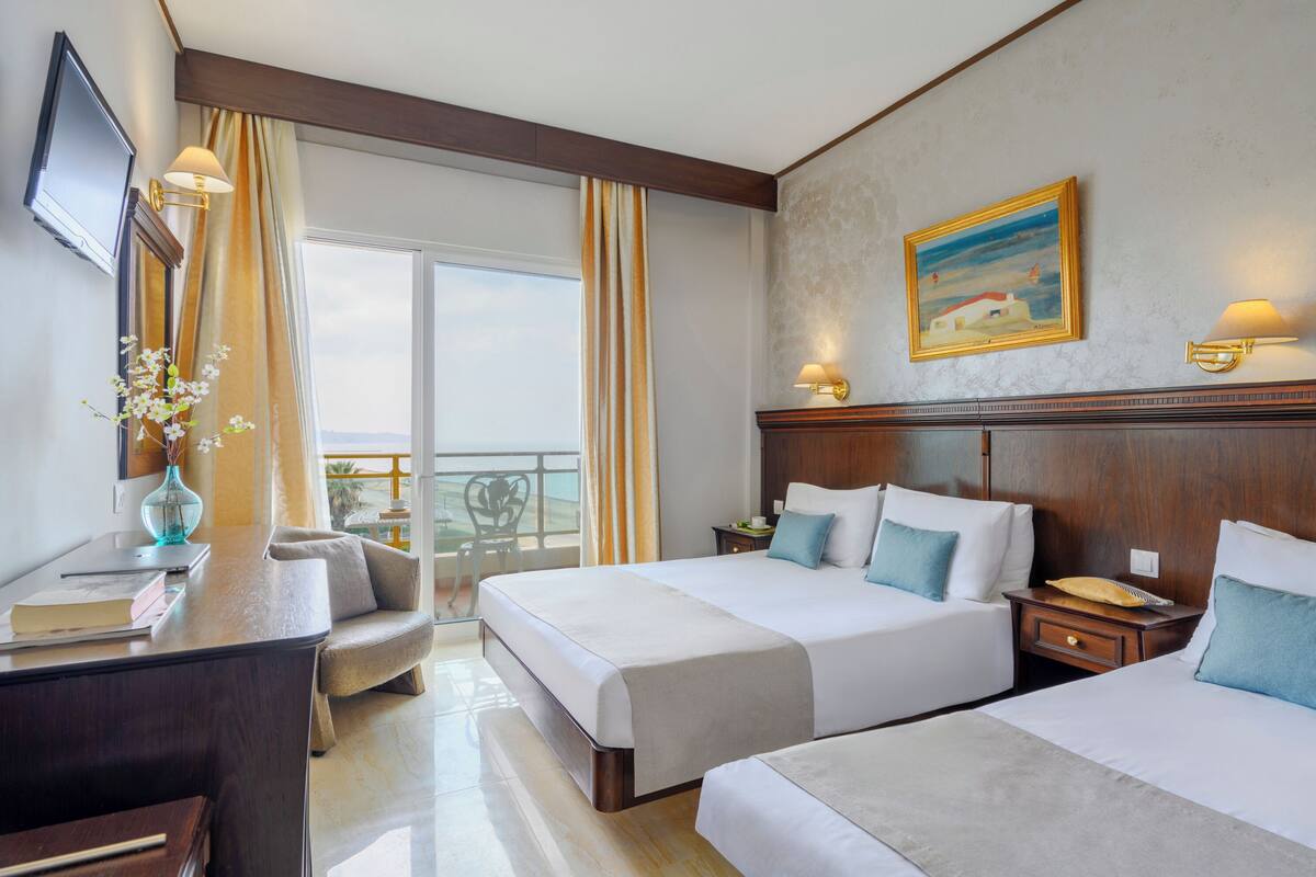 A hotel room with two beds, a desk, a chair, a TV, and a balcony with curtains.