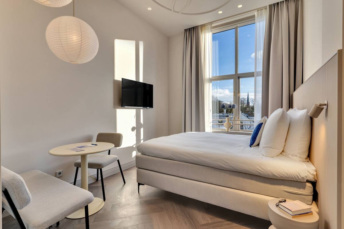 Ein modernes Hotelzimmer mit einem großen Bett, einem Fernseher, einem kleinen Tisch und Blick auf die Stadt.