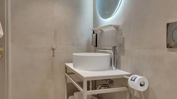 A modern bathroom with a white sink, toilet, and a mirror with a circular light. There is a shelf with towels and toiletries.