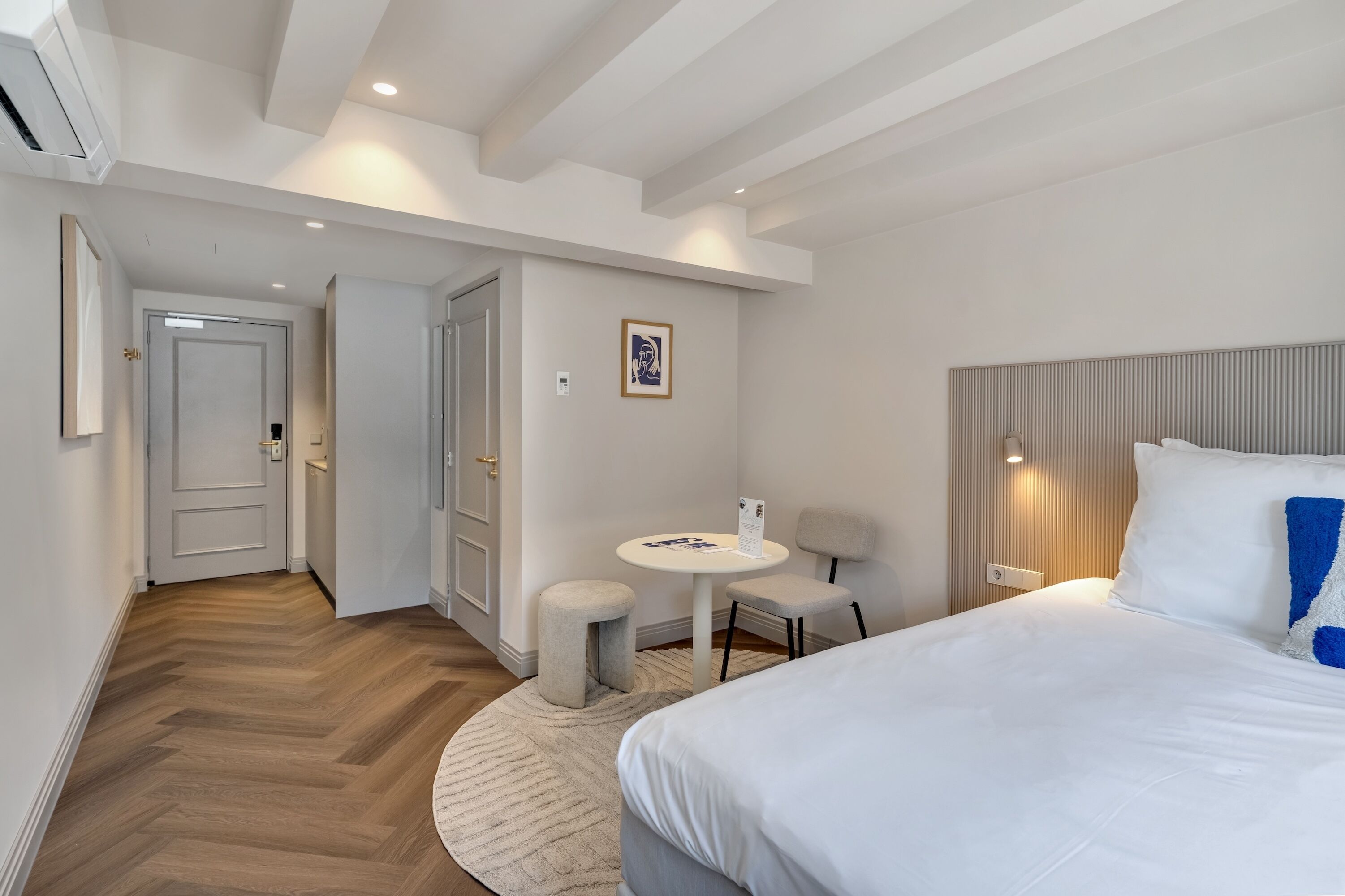 Una habitación de hotel moderna con una cama grande, una mesita redonda pequeña y una silla. Se ve una puerta que da a otra habitación y un aire acondicionado instalado en la pared.