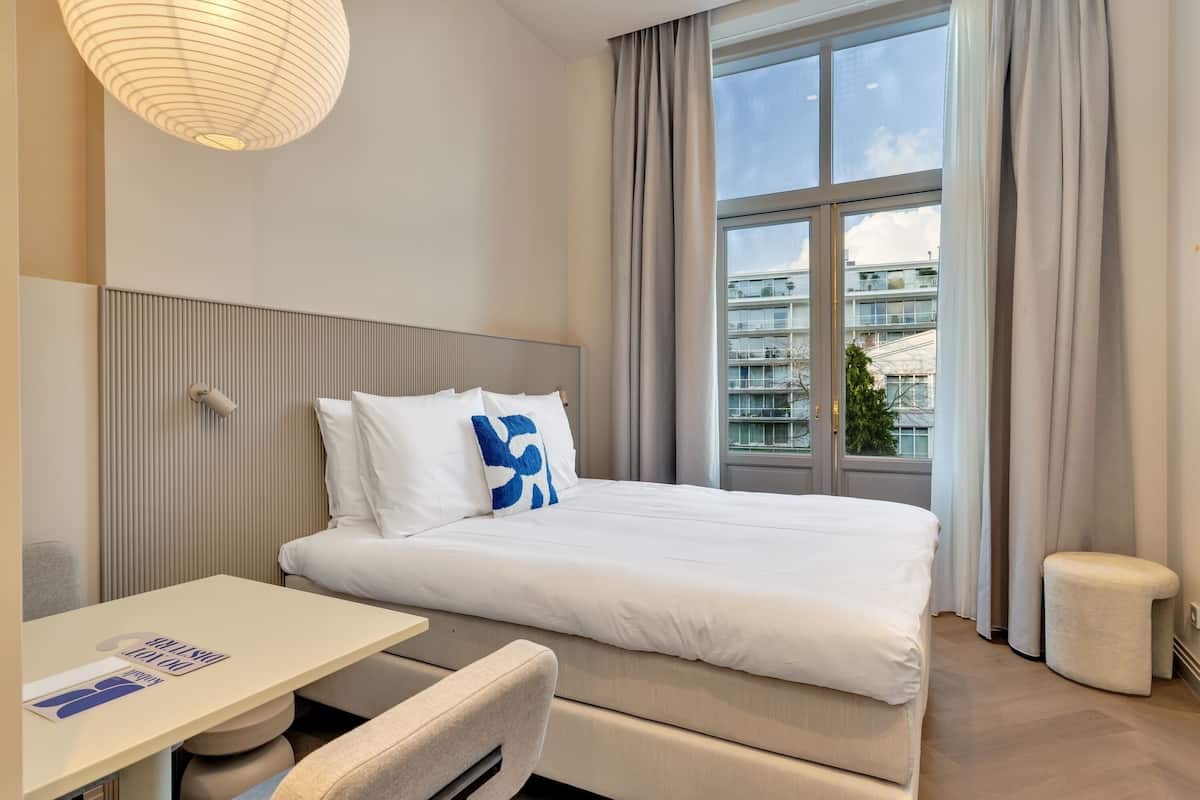 Ein modernes Hotelzimmer mit einem großen Bett, einem kleinen Tisch und einem Fenster mit Vorhängen.