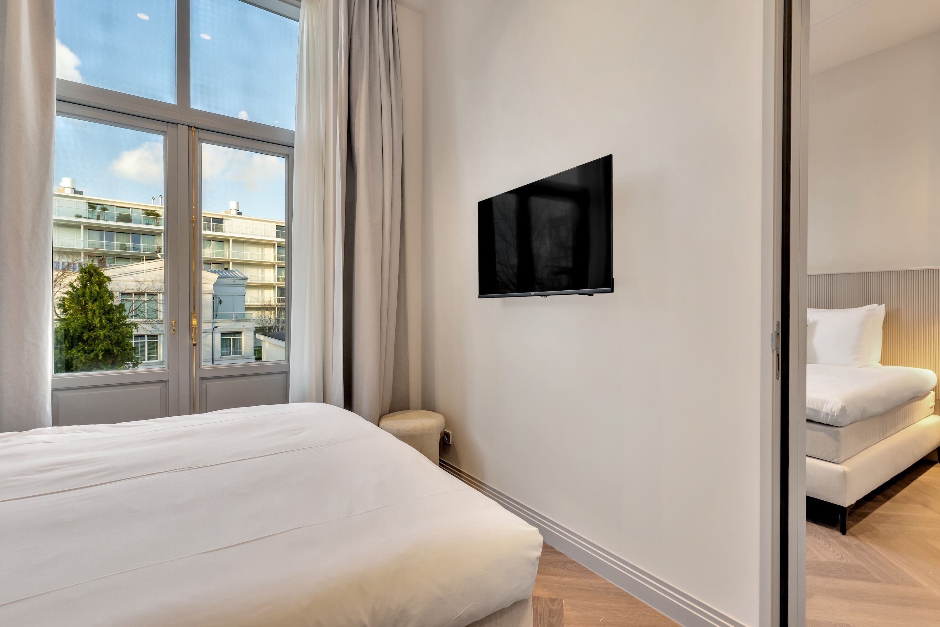 A modern hotel room with a large window, a flat-screen TV, a bed, and a sofa.
