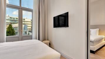 A modern hotel room with a large window, a flat-screen TV, a bed, and a sofa.