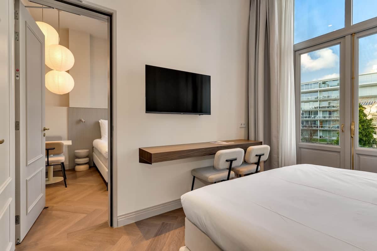 Ein modernes Hotelzimmer mit einem Bett, einem Schreibtisch mit Stühlen, einem Wand-Fernseher und einem großen Fenster mit Blick auf die Stadt.