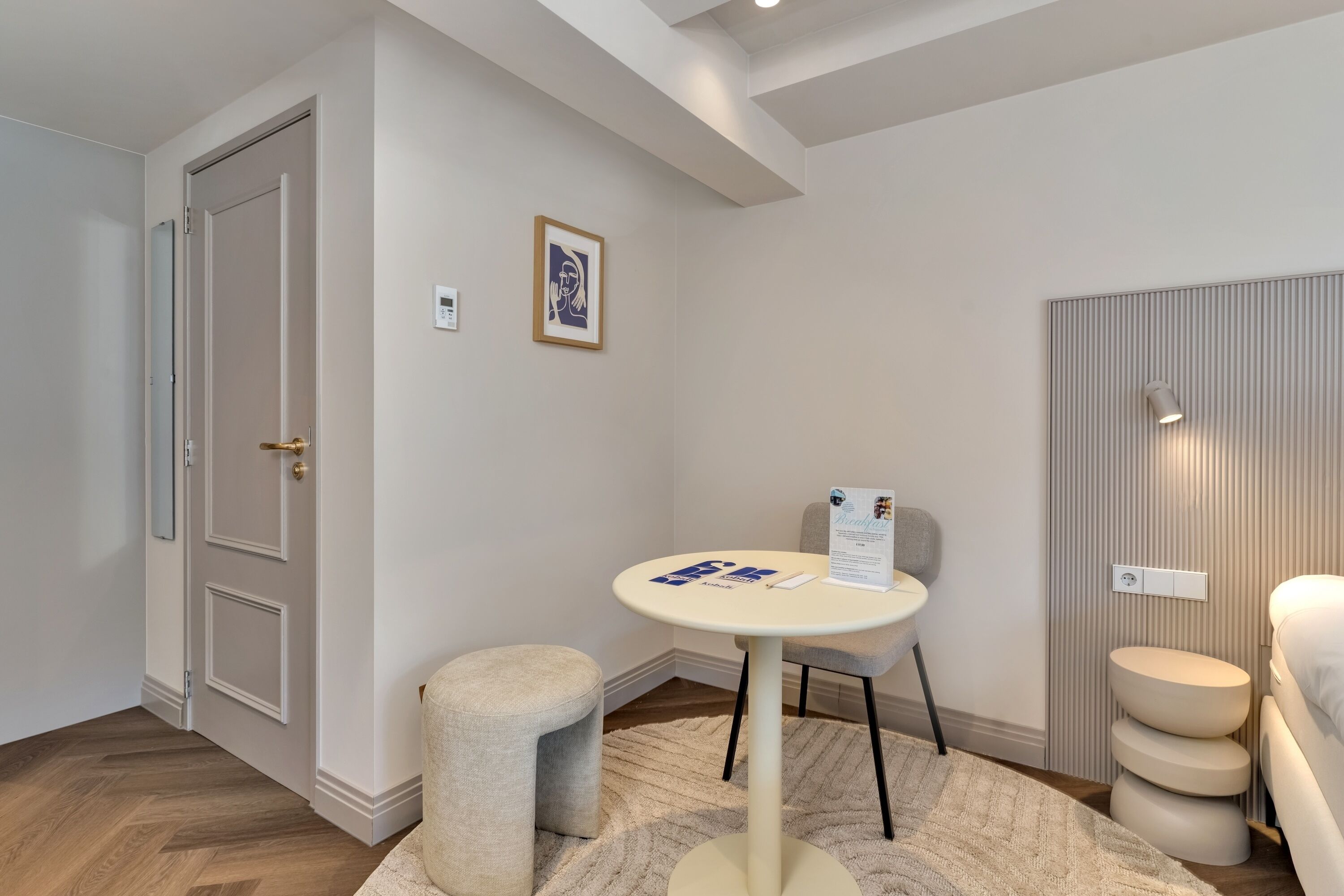 Quarto de hotel moderno com piso de madeira, uma mesa redonda com uma cadeira, um banquinho pequeno, uma cama com cabeceira e um quadro emoldurado na parede.
