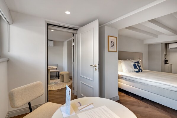 Una habitación de hotel moderna con una cama, un escritorio con silla, una mesita y una puerta que da a otra habitación.