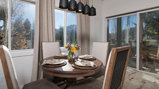 A dining area with a round table, chairs, a vase with flowers, and a view of trees through large windows. - Condo with Fireplace and Mountain Views! Close to Sun Valley Village! (Sun Valley)