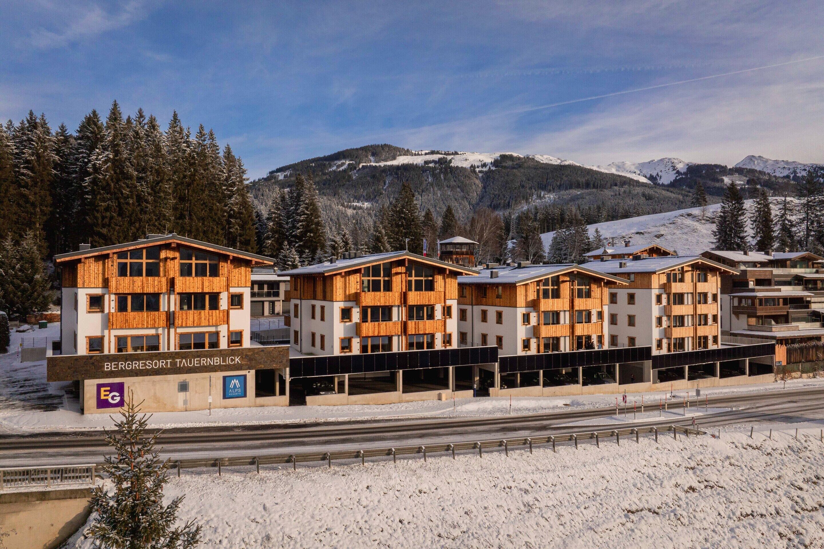 Un hotel a più piani con facciata in legno, situato vicino a una linea ferroviaria, circondato da montagne innevate.