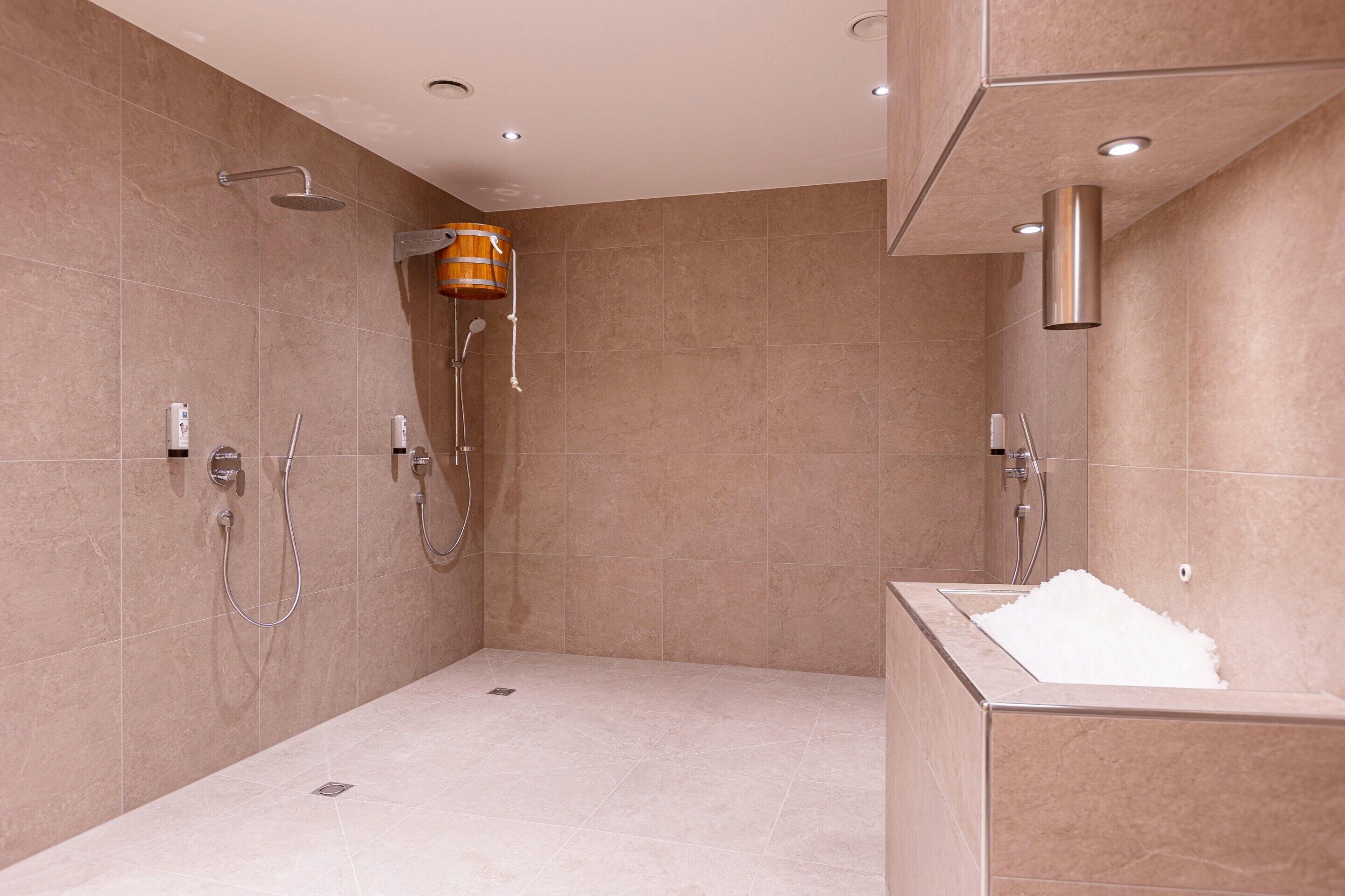 Una ducha moderna con un cubo de madera, un cabezal de ducha independiente y una bañera separada con una alfombra de baño blanca.