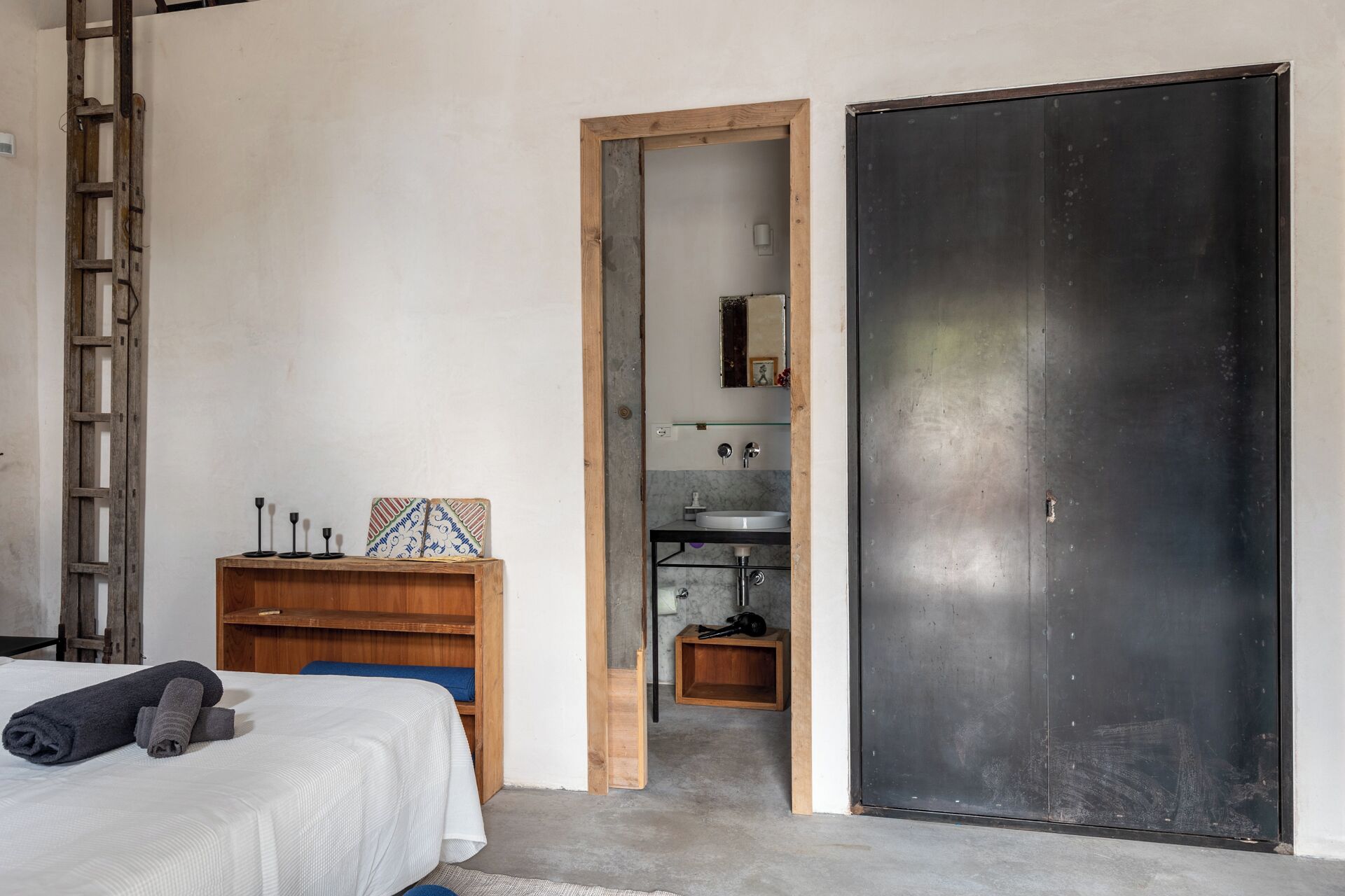 Une chambre à coucher avec un lit, une table de chevet en bois, une commode et une salle de bain visible à travers une porte ouverte.