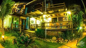 A two-story wooden house with a balcony, outdoor seating area, and a well-lit garden at night. - Pousada Canto da Vigia (Penha)