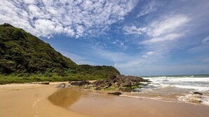 A beach with a sandy shore, rocky outcrops, and a forested hill in the background. - Pousada Canto da Vigia (Penha)