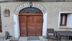 Eine hölzerne Doppeltür mit Glasfenster, eine kleine Bank und ein Tisch vor einem Gebäude mit einem Steinbogen und einem Fenster mit Holzklappe.