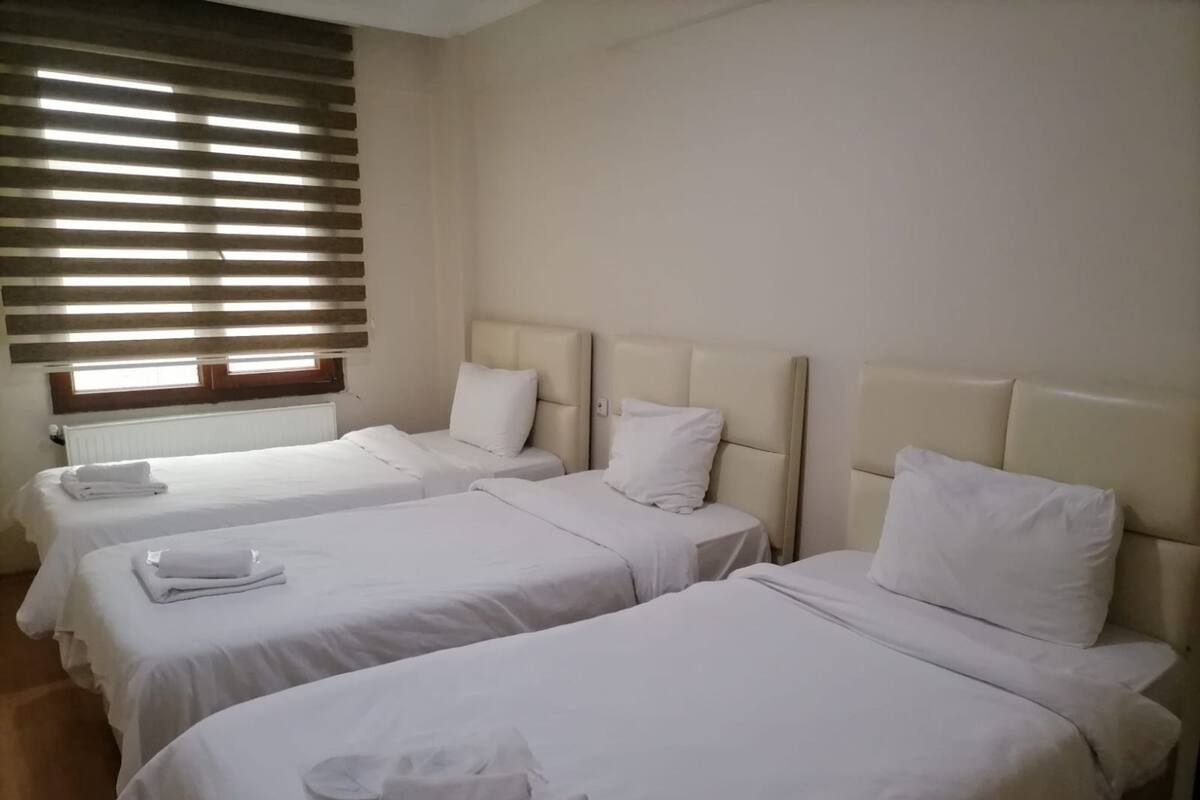 Une chambre d’hôtel avec trois lits simples, chacun doté d’une tête de lit et d’un oreiller blancs, et une fenêtre avec des stores horizontaux.