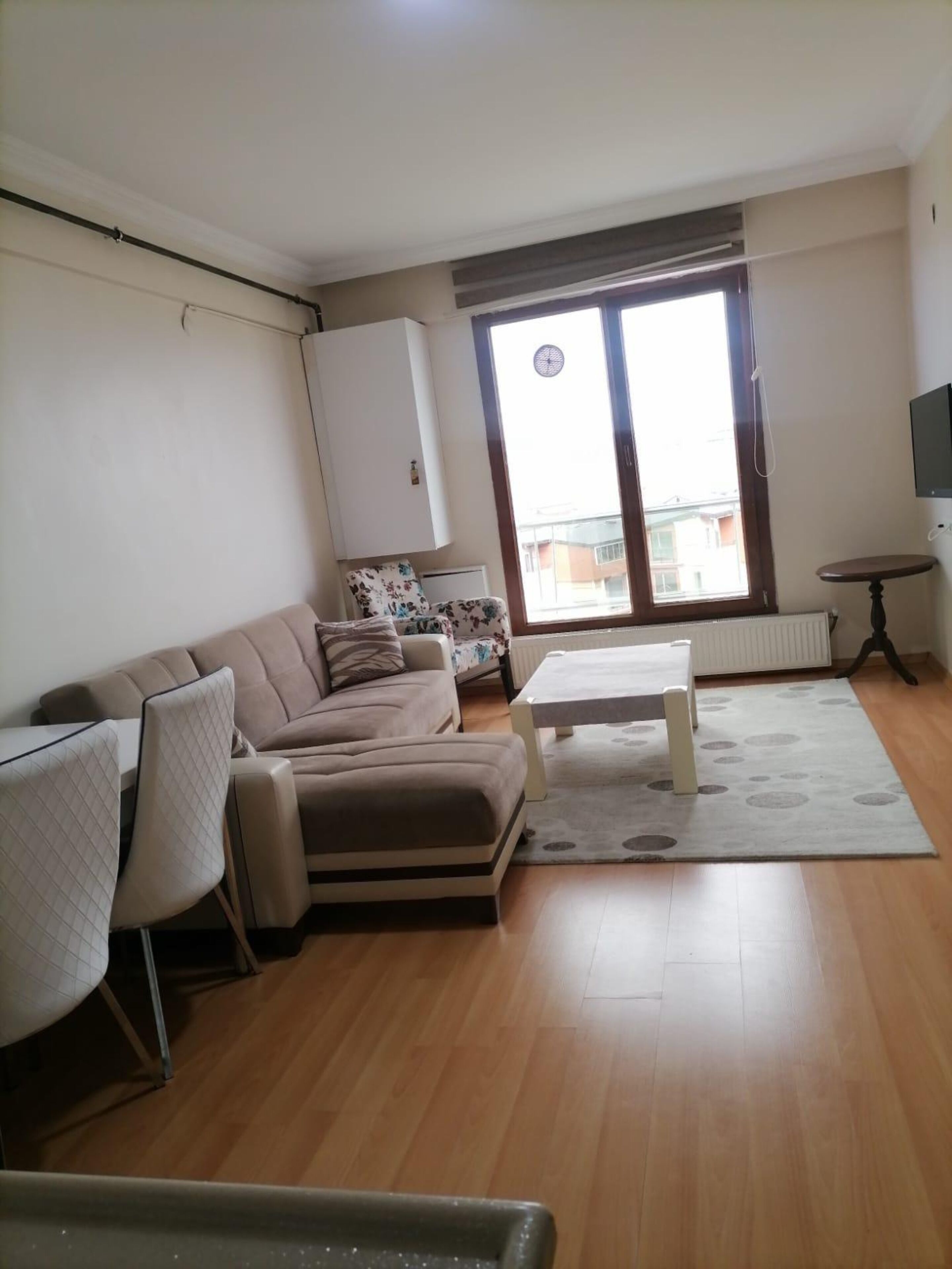 Una sala de estar con un sofá, una mesa de centro y una silla. Se ve una ventana con vistas a edificios.