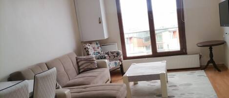 A living room with a sofa, a coffee table, and a chair. There is a window with a view of buildings outside.
