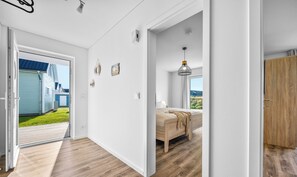 Un interior moderno con piso de madera, una cama con manta color beige, una lámpara colgante y vista a una casa azul a través de una puerta de vidrio.