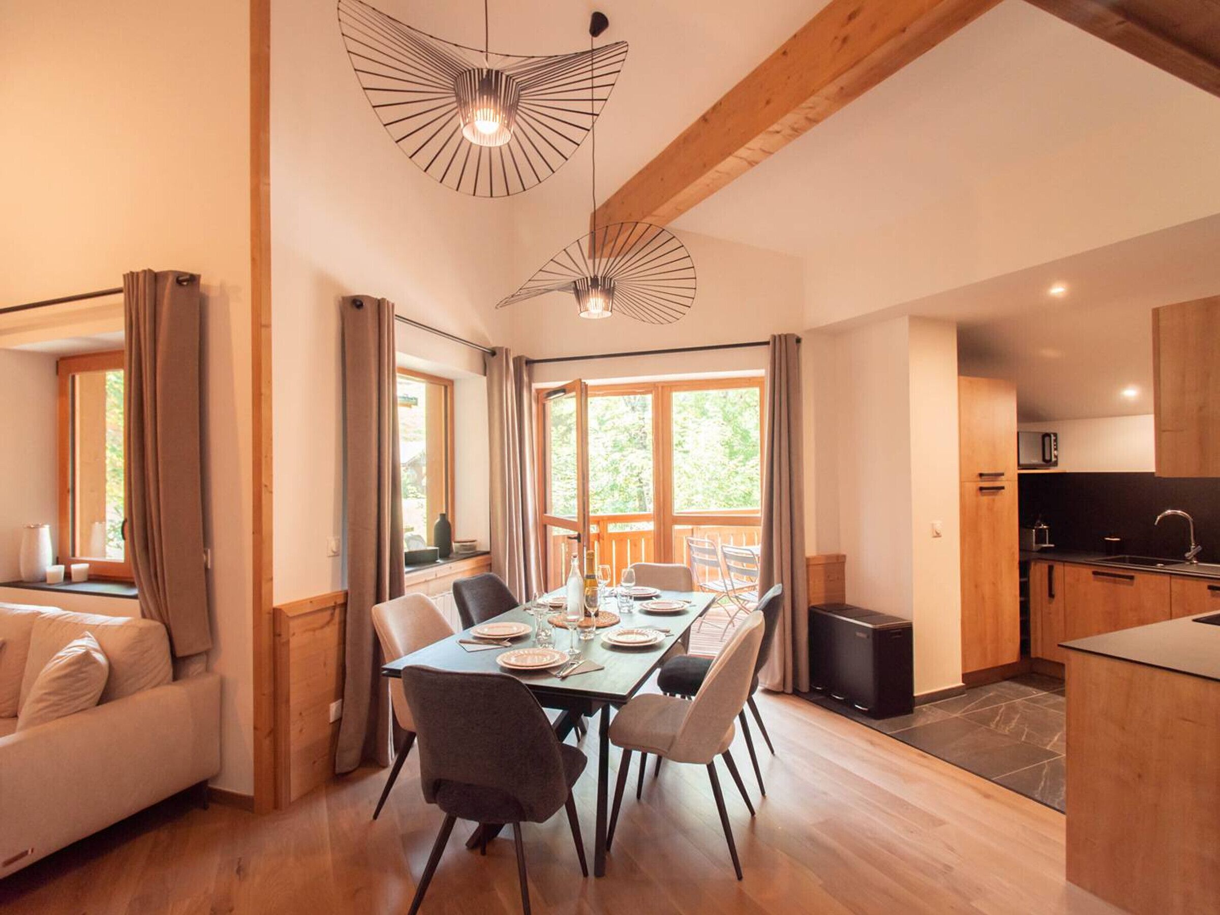 A modern kitchen with wooden cabinets, a dining area with a table set for four, and large windows providing natural light.