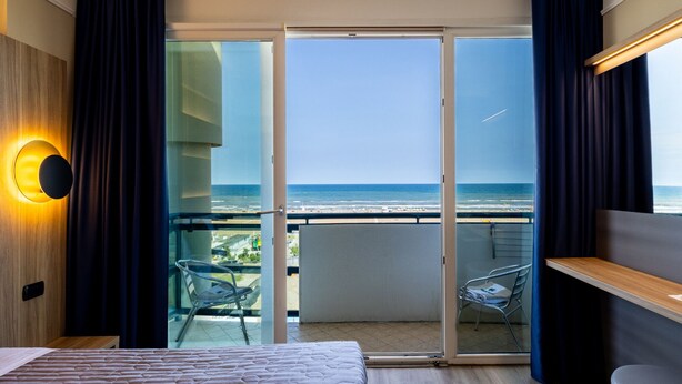 A modern hotel room with a large bed, a balcony with a glass door, and a view of the ocean. - Hotel Palm Beach (Rimini)