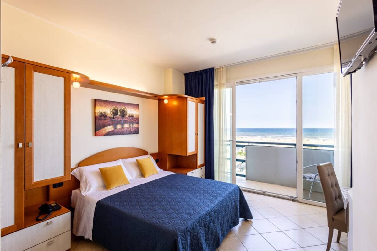 A hotel room with a bed, a desk, a chair, a painting, and a balcony with a view of the beach.