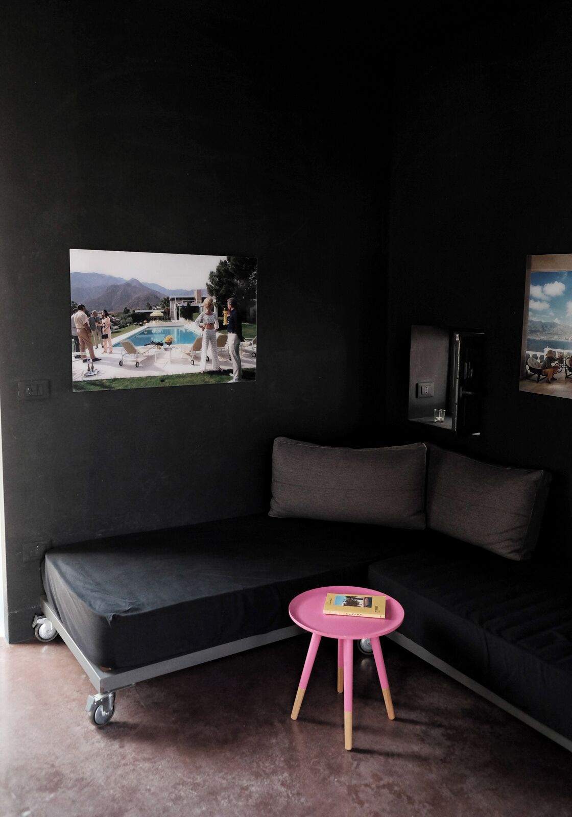 Ein dunkles Zimmer mit einer Couch, einem kleinen rosafarbenen Tisch und zwei an der Wand befestigten Fernsehern.