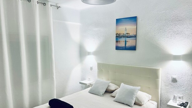 A hotel room with a large bed, white bedding, a headboard, two bedside tables with lamps, a painting of a lighthouse, and a curtain. - Aperol Apartment - Malaga (Málaga)