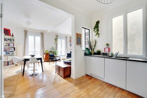 A modern kitchen with white cabinets, a wooden dining table, and a large window. - Martyrs Village Montmartre 2 habitaciones (París)