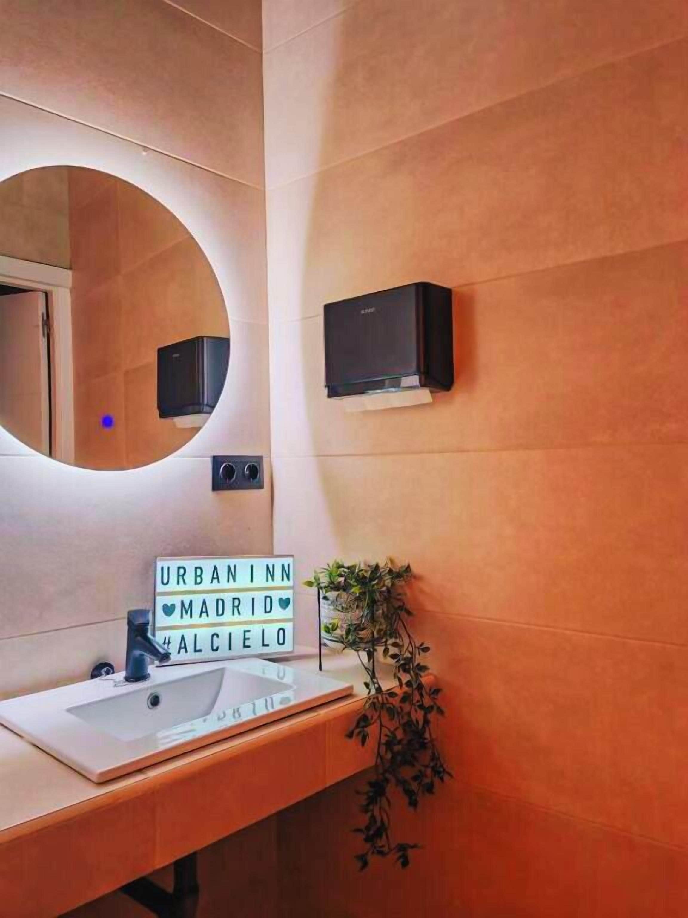Un baño con un espejo redondo, un lavamanos, un cartel que dice "URBAN INN MADRID MALCUELO", un pequeño altavoz negro y una planta en maceta.
