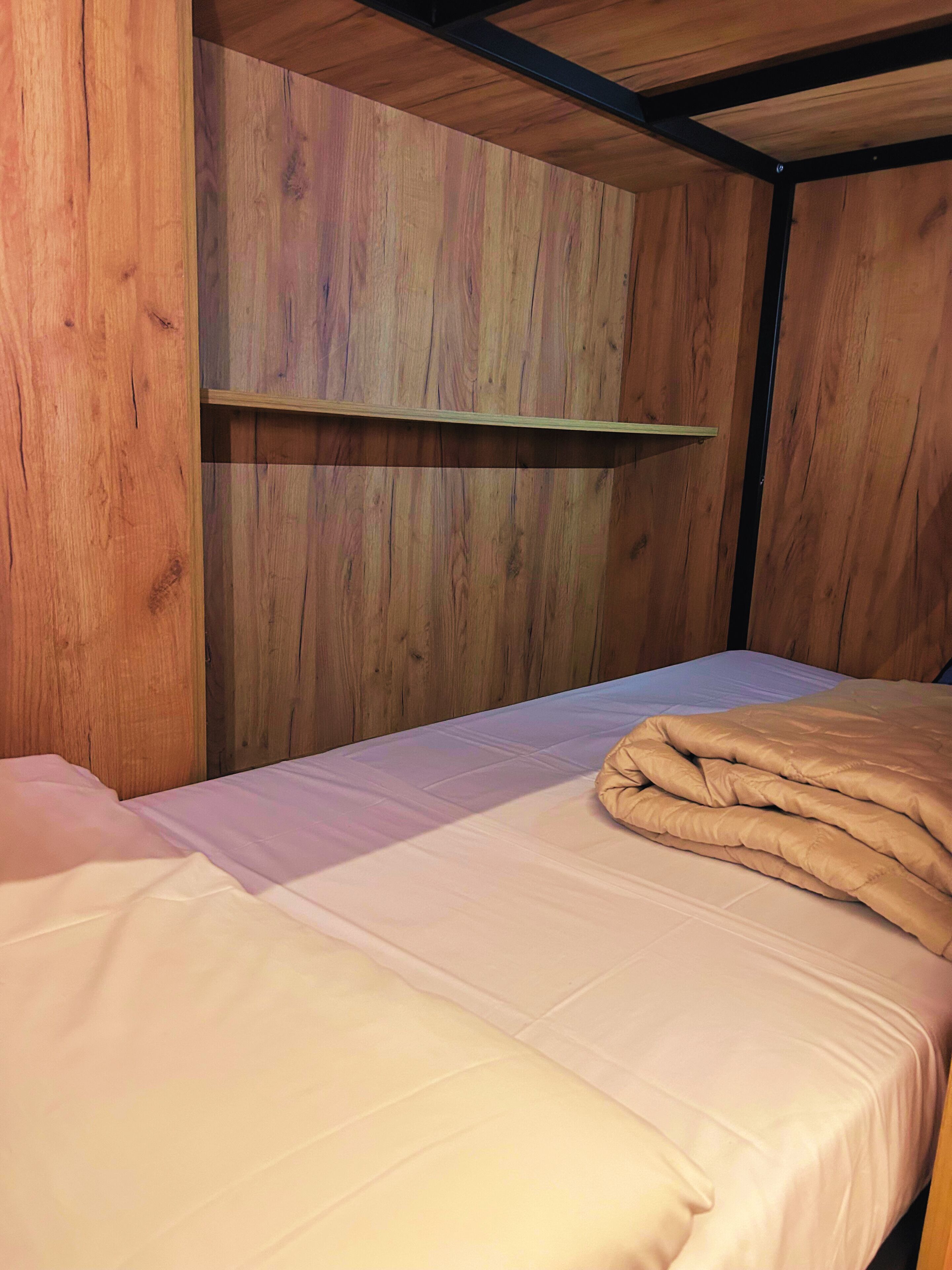 A single bed with a white mattress and a beige blanket, wooden walls, and a simple shelf unit.