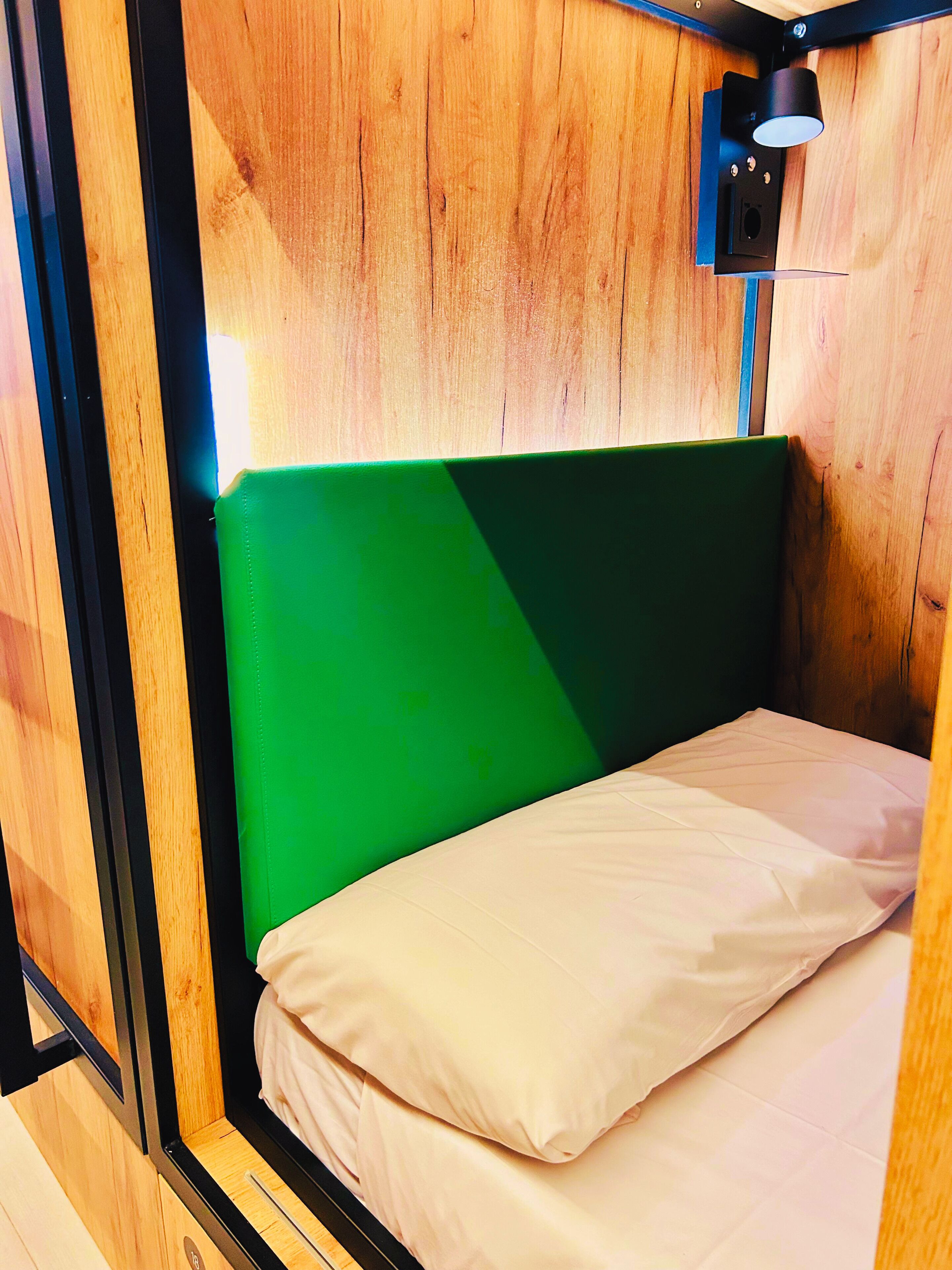 A small, compact room with a green headboard, a single bed with white bedding, and a small wall-mounted light fixture.