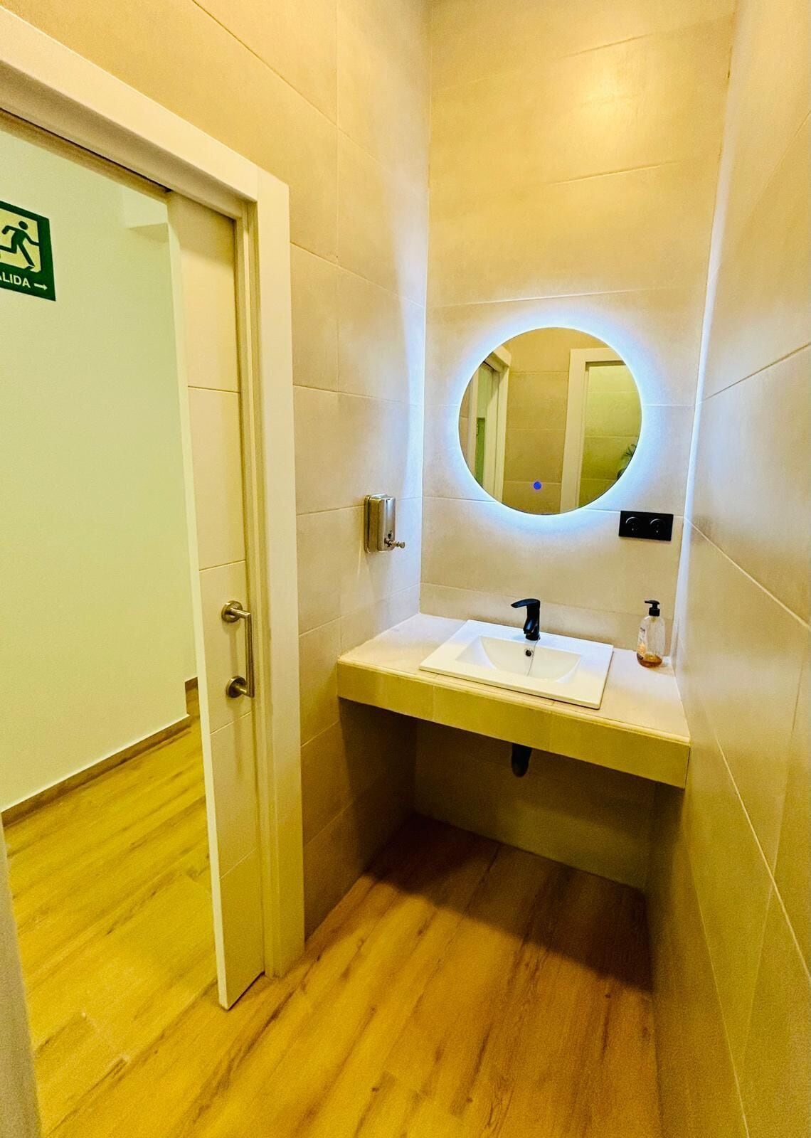 A small bathroom with a white sink, a round mirror, and a door with a green sign.
