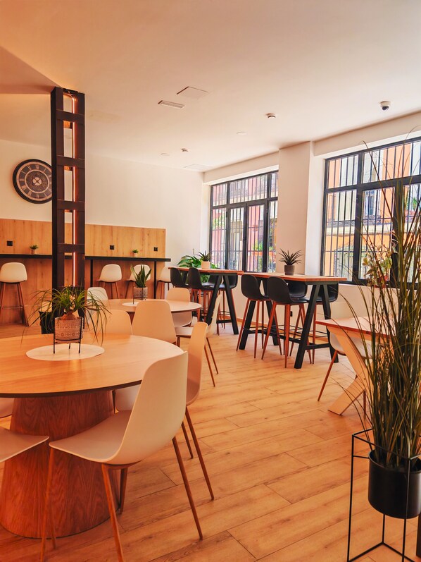A modern dining area with wooden tables and chairs, potted plants, and large windows offering views of the outside. - Capsule Inn Madrid (Madrid)