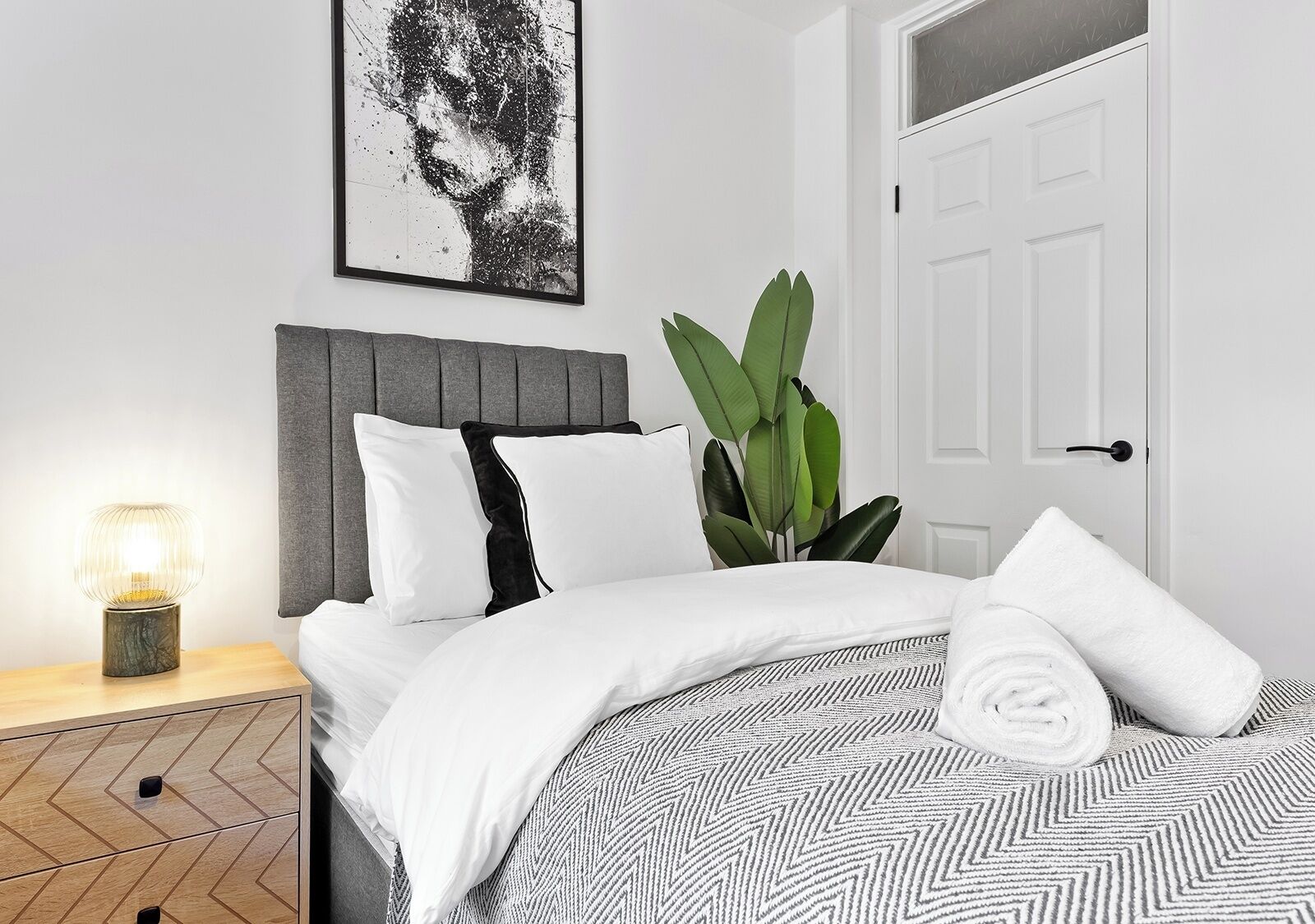 A neatly made bed with white bedding and a patterned throw, a grey upholstered headboard, a potted plant, and a framed artwork on the wall.