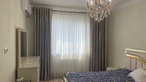 A bedroom with a bed, a desk, a chair, a mirror, a chandelier, and a window with curtains. - Chaqar 317 (Tashkent)
