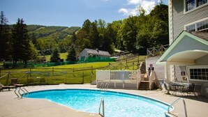 Una piscina con solarium, zona salotto coperta e vista su una casa verde e colline boscose.