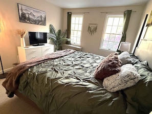 Un dormitorio con una cama, un televisor en una cómoda, una ventana con cortinas y una planta.
