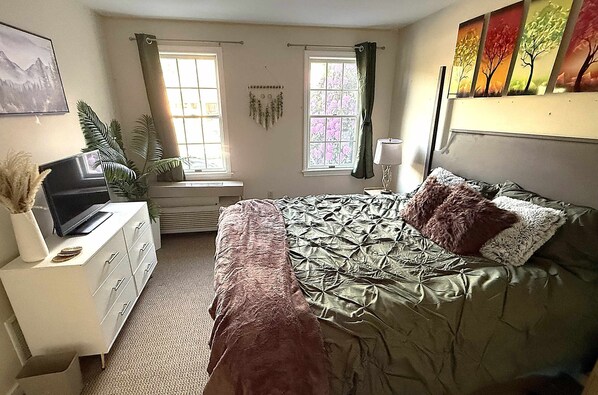 Un dormitorio con cama, cómoda, televisor y dos ventanas con cortinas.