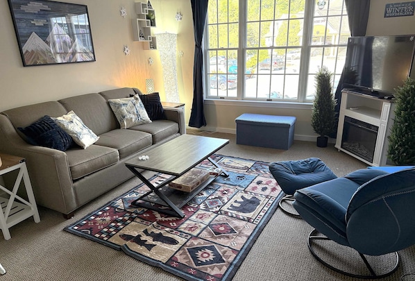 Una sala de estar con sofá, mesa de centro, reposapiés, alfombra estampada, televisor y ventana con vistas al exterior.