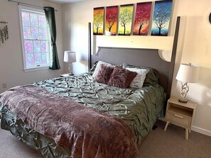 Ein Schlafzimmer mit einem Bett, einem Nachttisch, einer Lampe, einem Fenster mit Vorhängen und einer Wand mit mehreren Bildern.